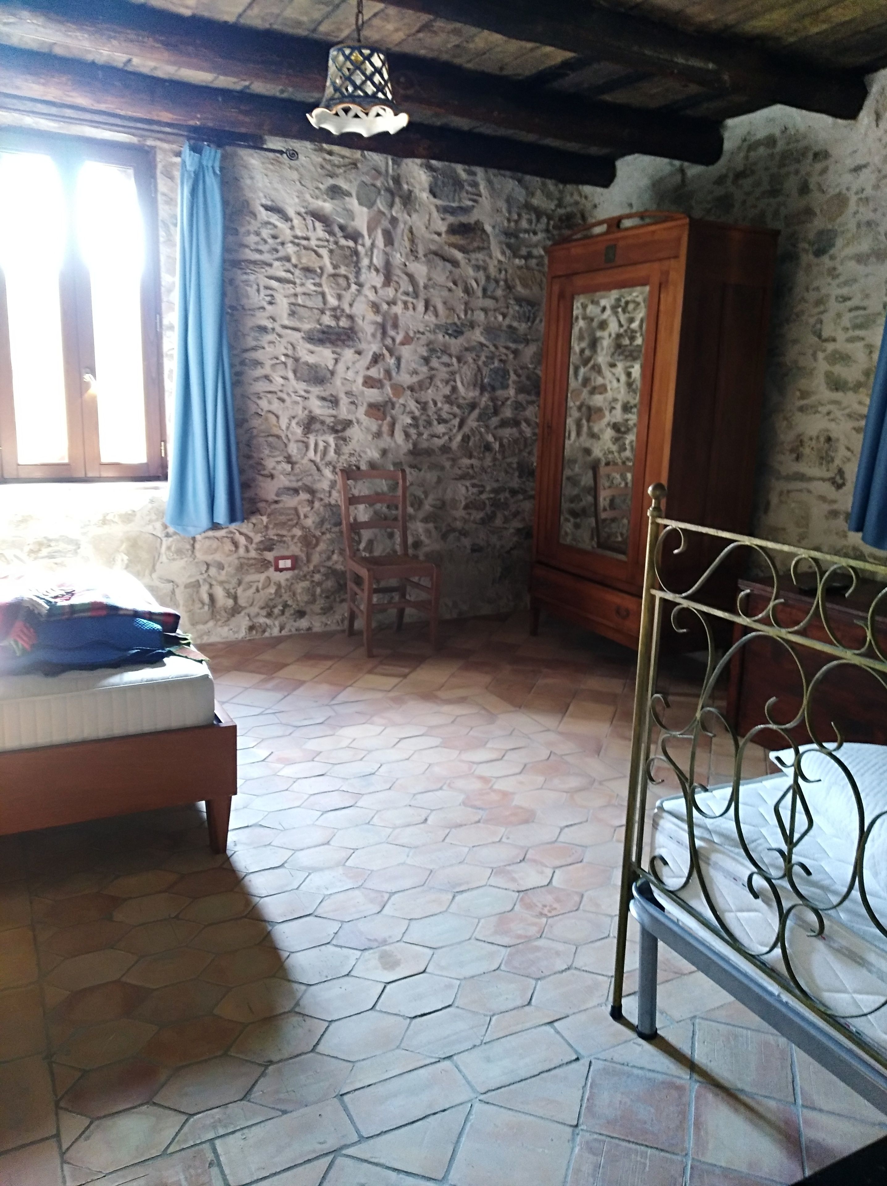 Quarto com parede de pedra, cama de madeira, cabeceira de metal, guarda-roupa de madeira e uma cadeira.