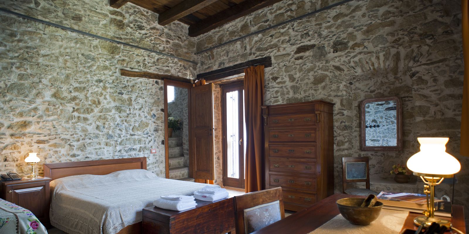 Un dormitorio rústico con pared de piedra, cama de madera, cómoda y una mesita de noche con lámpara.