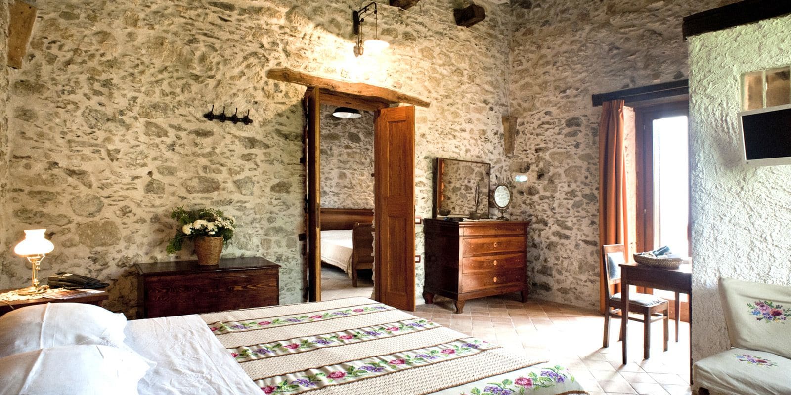 A rustic bedroom with a stone wall, a wooden bed, a dresser, and a chair.