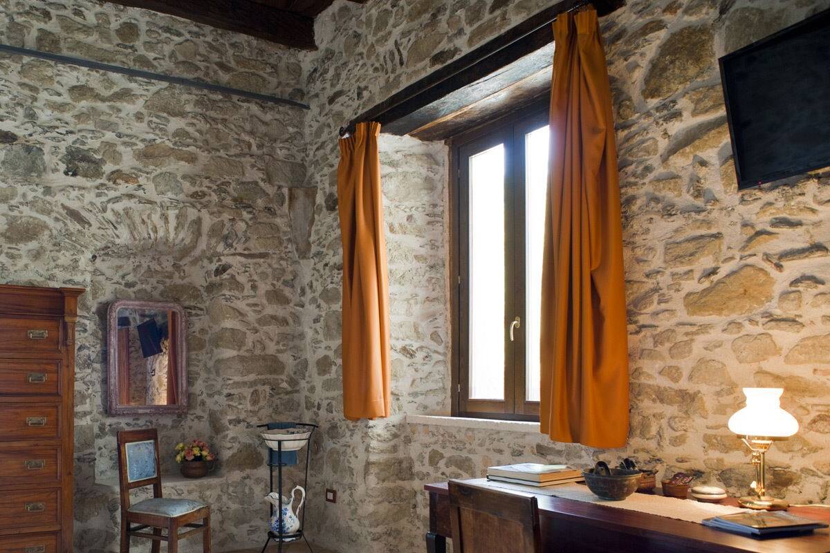 A rustic room with stone walls, a wooden dresser, a desk with a lamp, and a window with curtains.