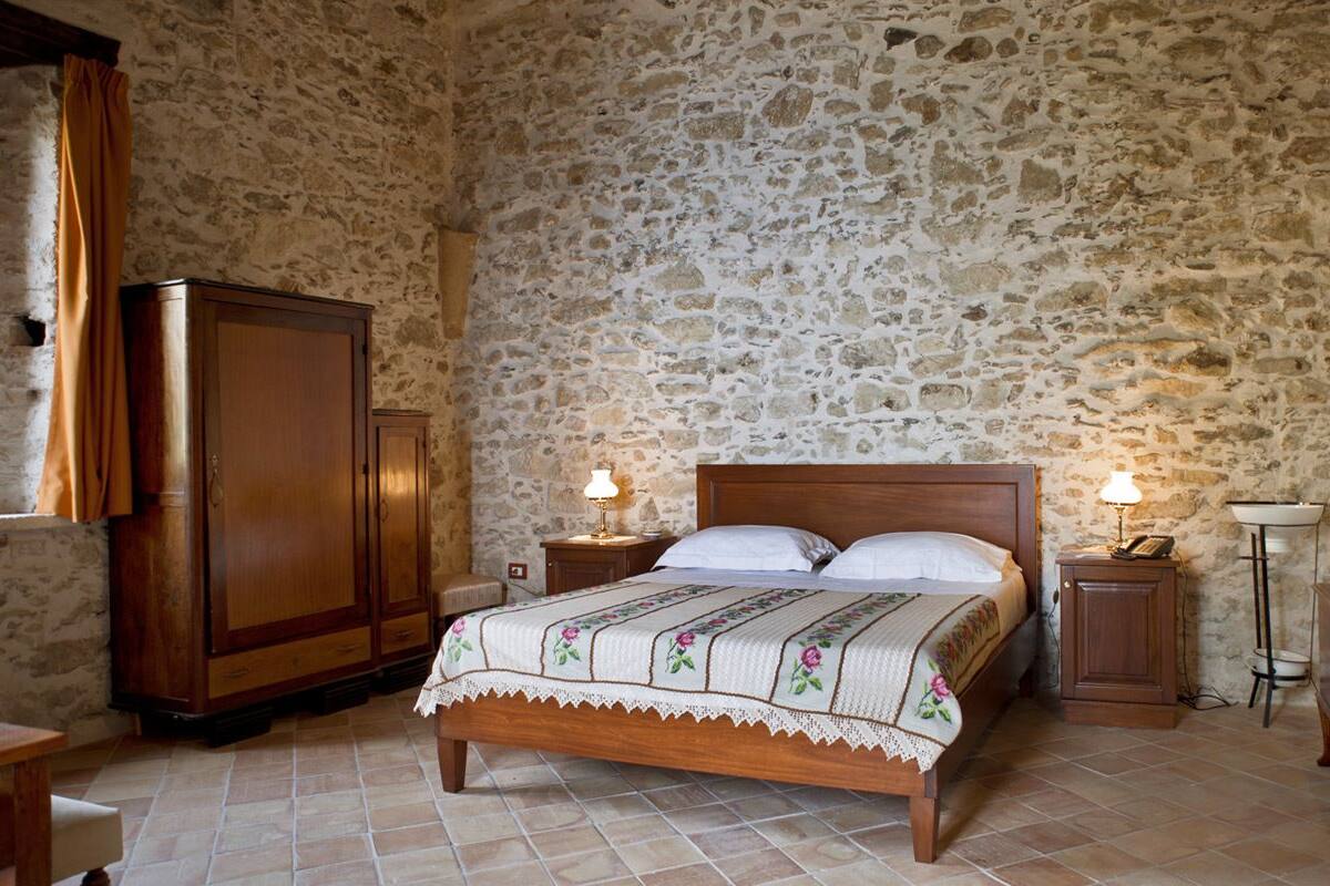 A bedroom with a stone wall, a wooden bed, a nightstand with a lamp, a wooden wardrobe, and a small table with a plant.
