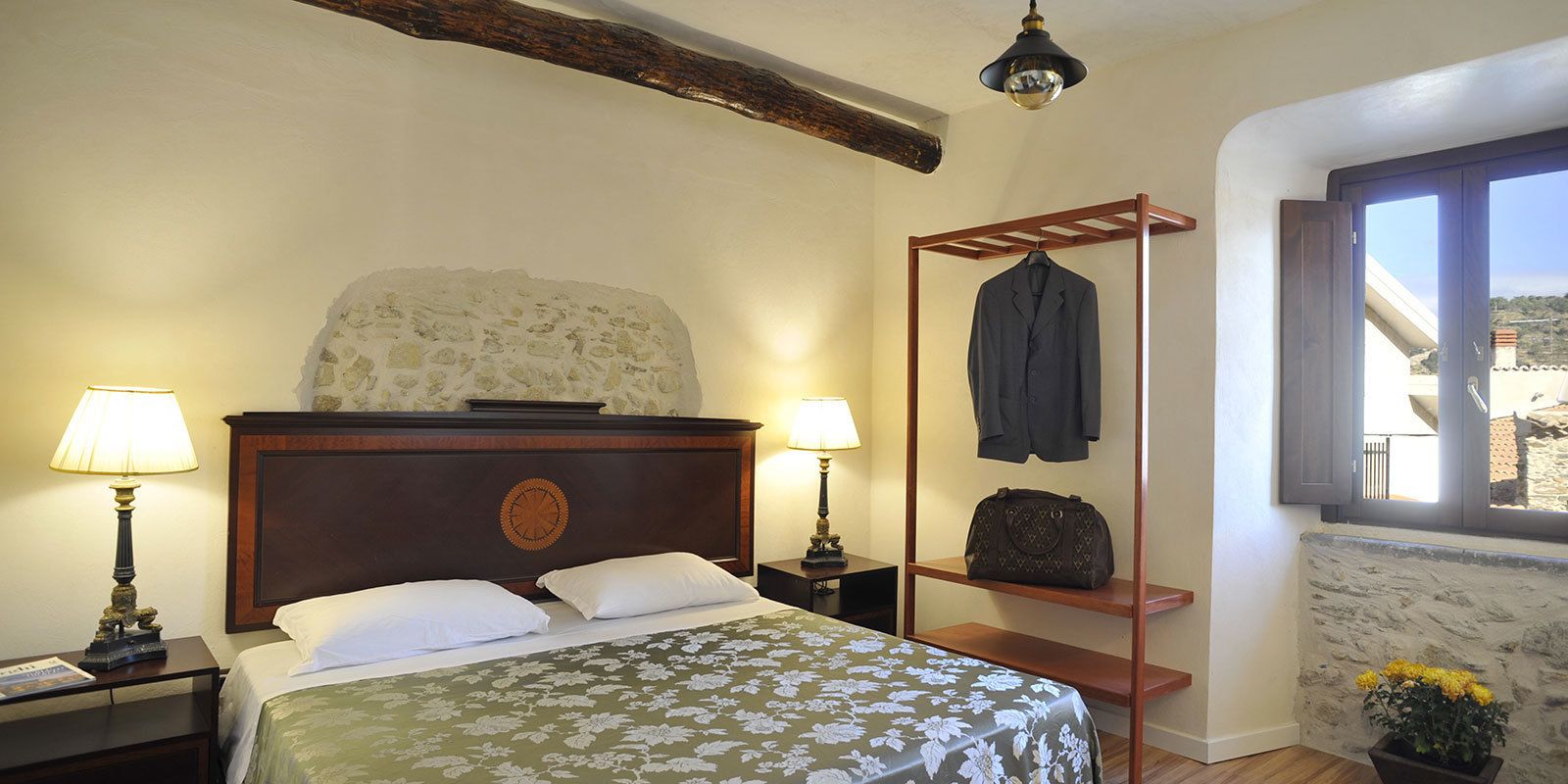 A hotel room with a bed, two bedside lamps, a wooden headboard, a hanging coat rack, a window with a view, and a potted plant.