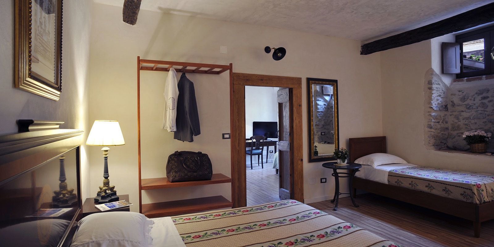 A bedroom with a bed, a bedside table with a lamp, a wooden shelf with clothes and a handbag, a small table with a plant, and a mirror on the wall.