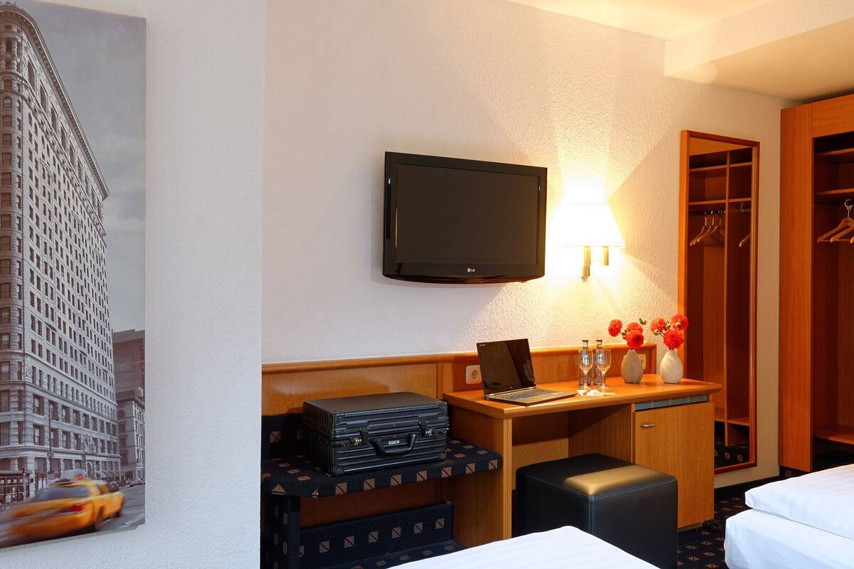 A hotel room with a bed, a desk with a laptop, a television, a wardrobe, and a painting of a building.