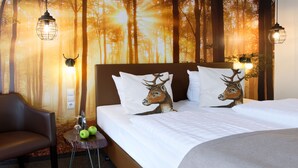 A bedroom with a forest-themed wall mural, a bed with deer pillows, a side table with apples and a bottle, and two hanging lights. - Trend Hotel Oldenburg (Wiefelstede)
