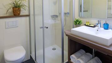 A bathroom with a glass-enclosed shower, a white toilet, a sink with a mirror, and a shelf with towels.