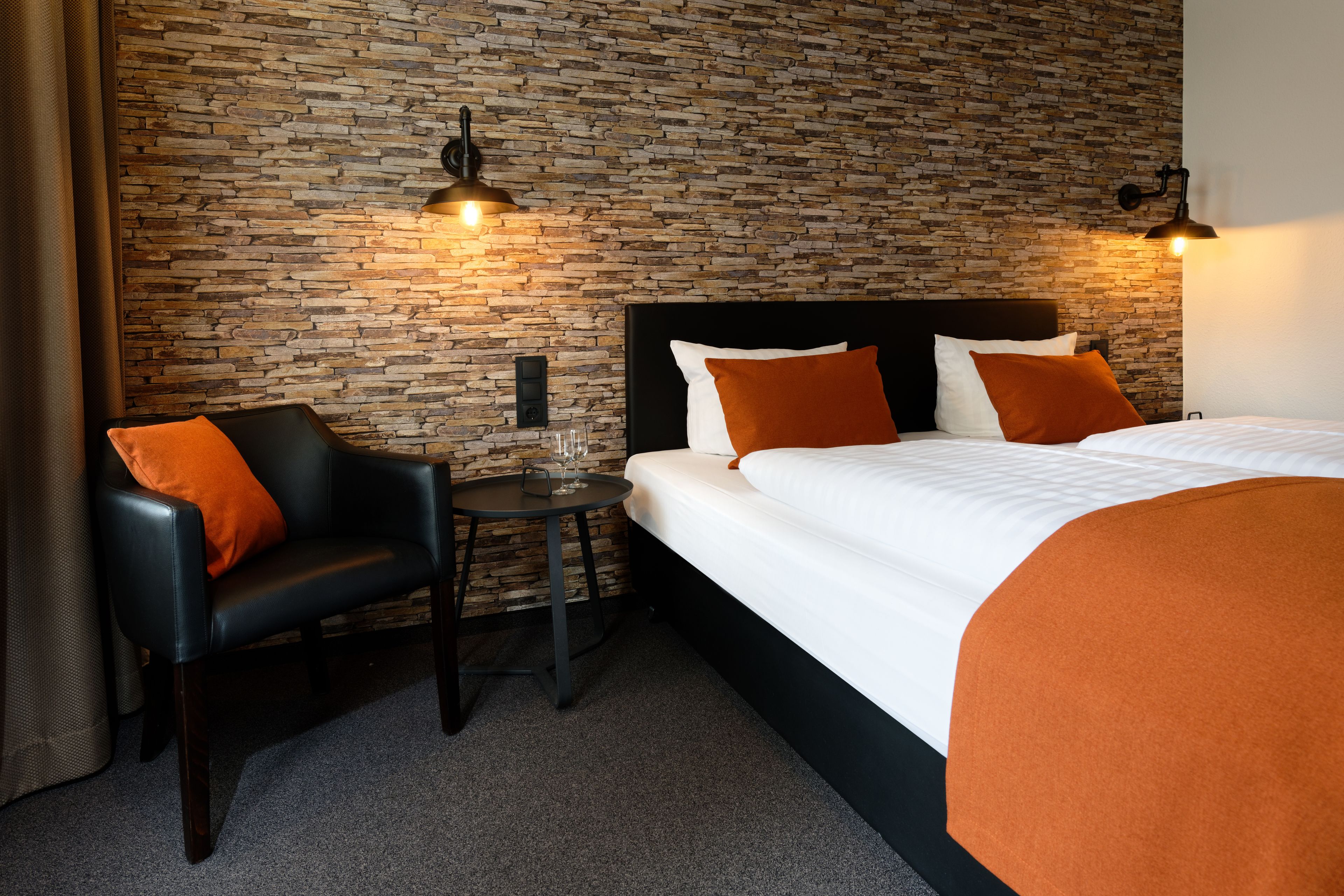 Ein Hotelzimmer mit einer Steinsäule, einem Bett mit weißer Bettwäsche und orangefarbenen Kissen, einem schwarzen Ledersessel und einem kleinen runden Tisch mit einem Glas.