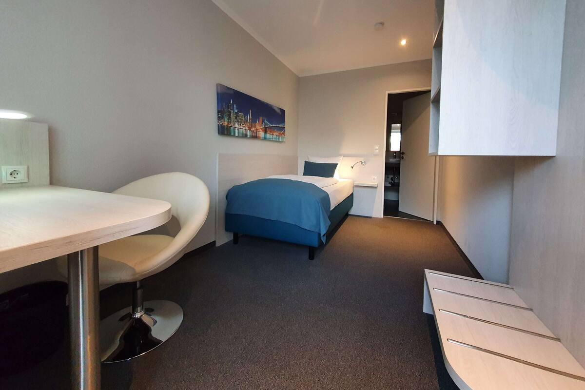 A hotel room with a bed, a desk, a chair, a painting, and a bathroom visible through an open door.