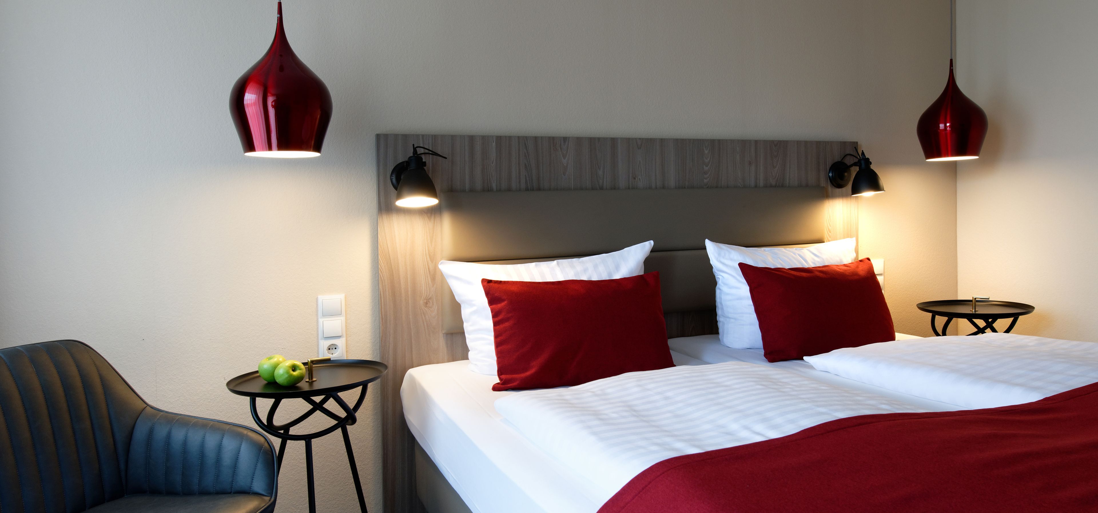 Een hotelkamer met een bed, twee rode lampen, een stoel en een kleine tafel met appels.