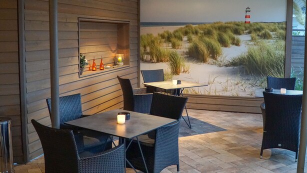 A cozy outdoor seating area with wicker chairs and tables, a wooden wall with a window, and a mural of a beach scene. - Trend Hotel Oldenburg (Wiefelstede)