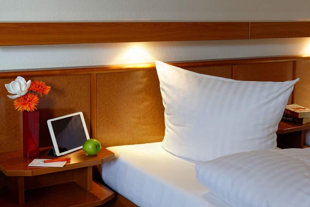 A hotel room with a wooden headboard, a bedside table with a tablet, a green apple, a red vase with flowers, and a white pillow.