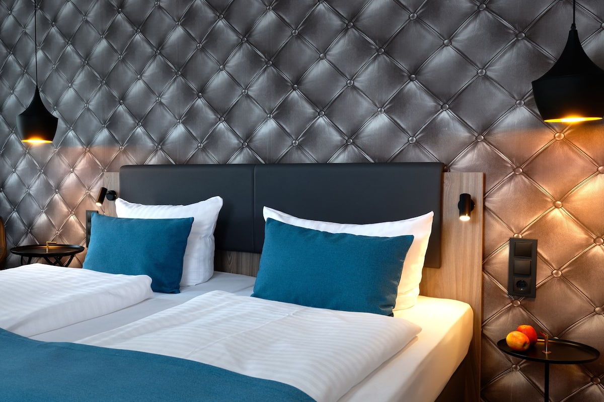 A hotel room with a tufted headboard, a bed with white and blue bedding, two bedside tables with lamps, and a small table with fruit.