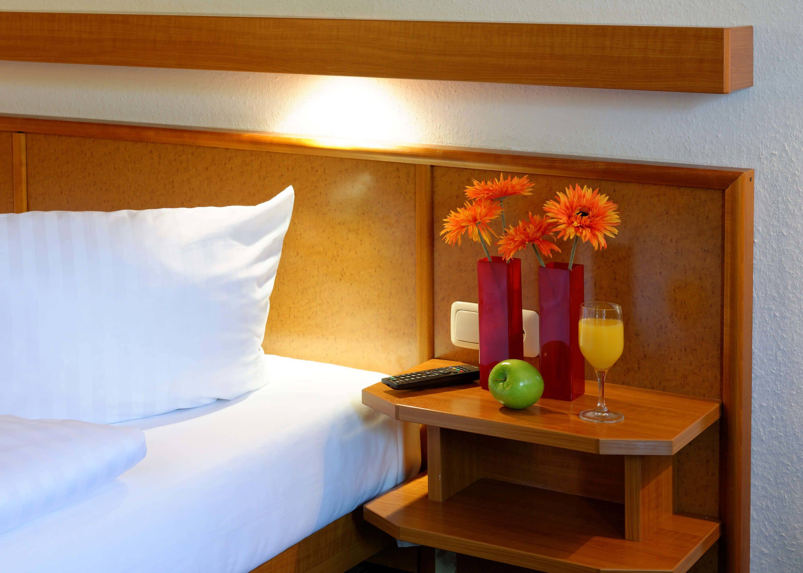A hotel room with a neatly made bed, a bedside table with a remote control, an apple, a glass of orange juice, and two red vases with orange flowers.