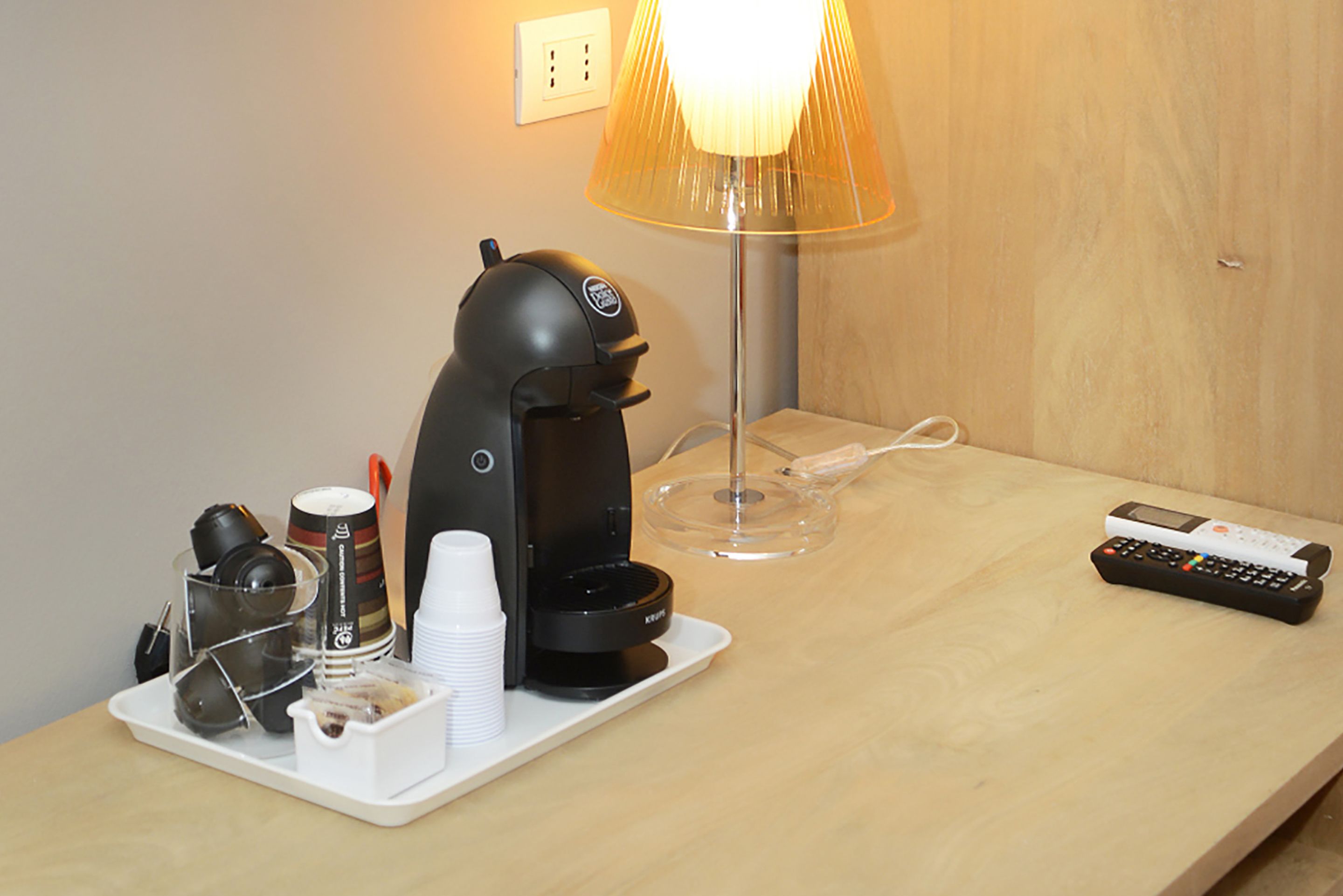 A coffee machine, cups, a remote control, and a lamp on a wooden table in a hotel room.