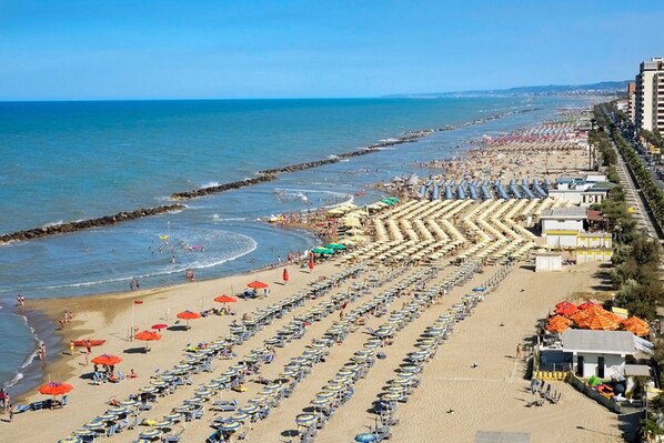 Una playa con hileras de sombrillas y tumbonas, un rompeolas rocoso y edificios a lo largo de la orilla.