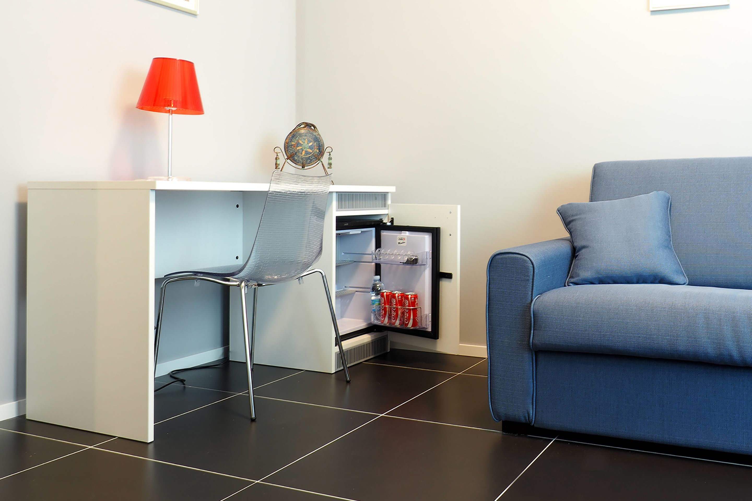A compact living space with a kitchenette, a blue sofa, a red lamp, and a chair.