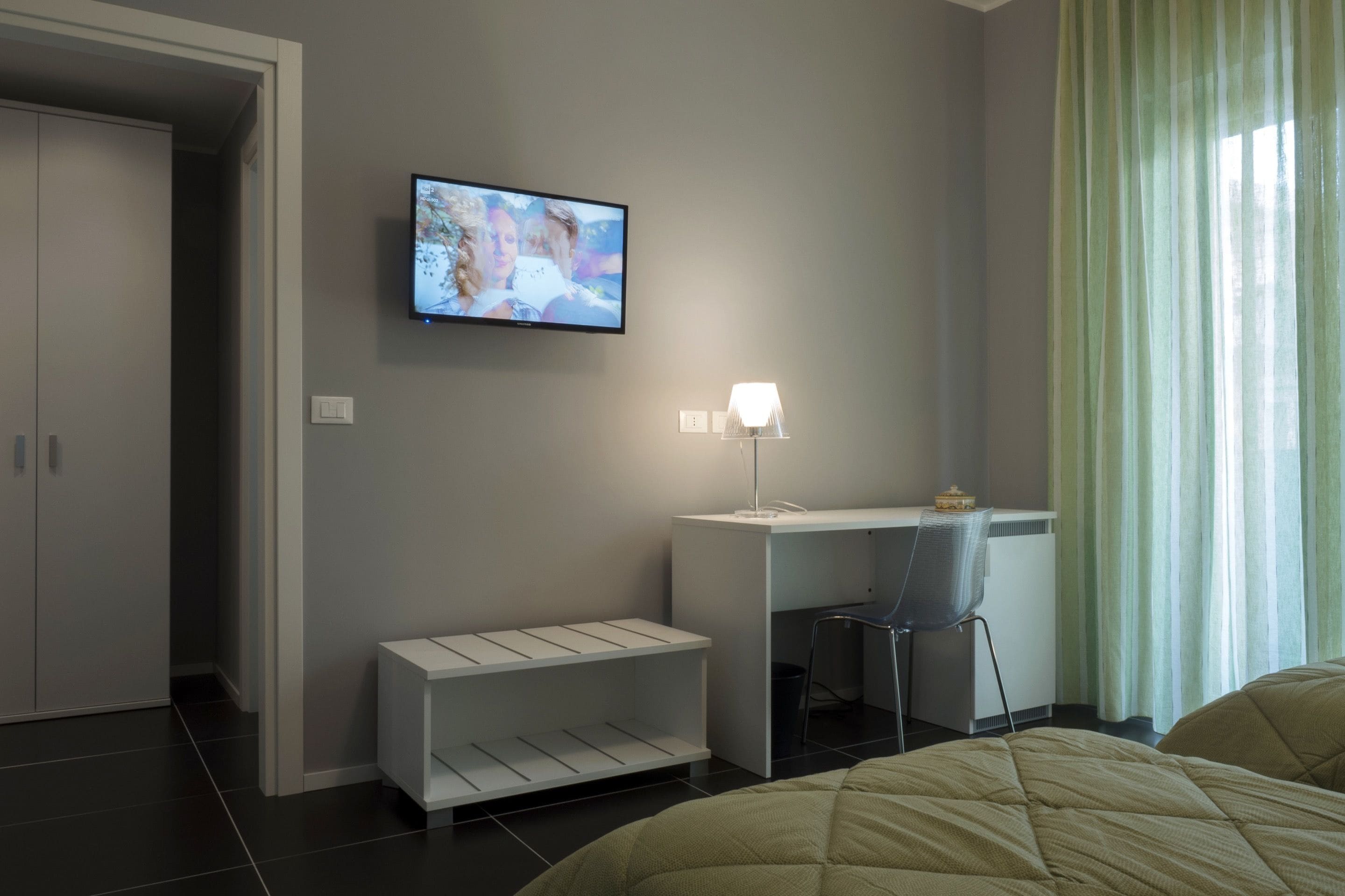 A modern hotel room with a bed, a desk, a chair, and a TV mounted on the wall.