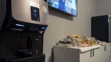 Una màquina de cafè amb pantalla digital i botons, una televisió penjada a la paret i una taula de buffet amb diferents plats.