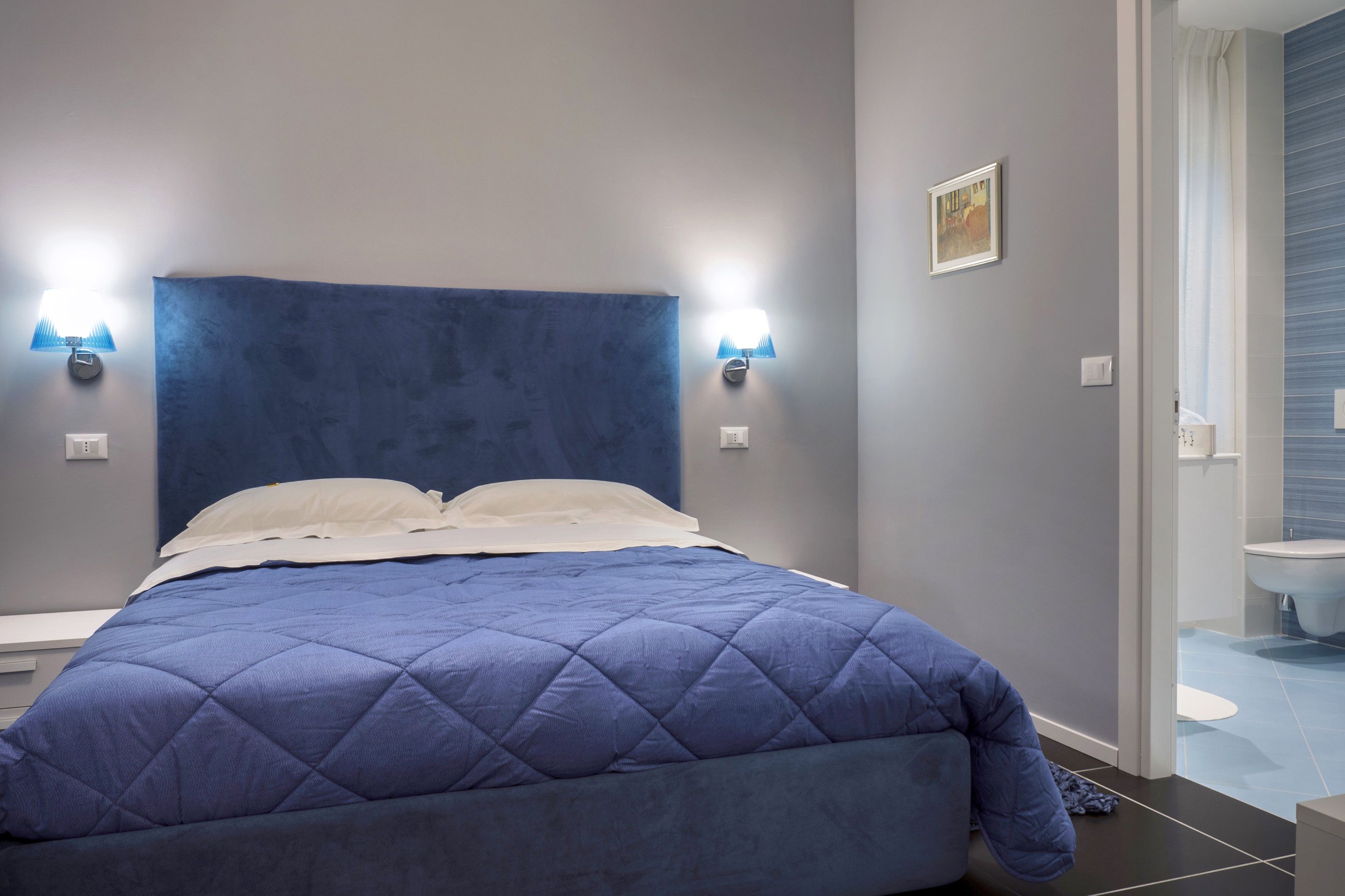 Une chambre moderne avec un grand lit, deux lampes fixées au mur et une salle de bain visible à travers une porte ouverte.