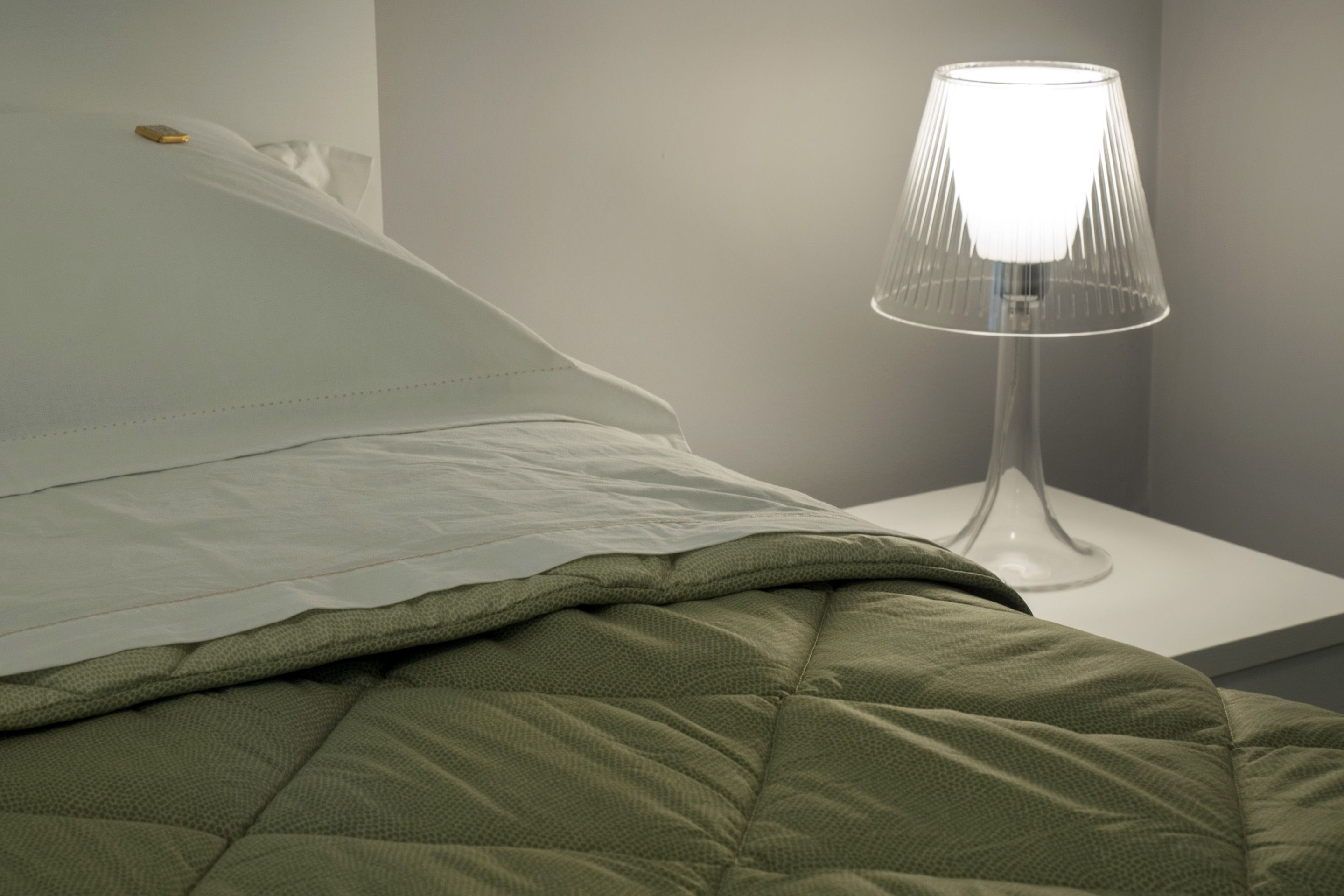 Een netjes opgemaakt bed met een groene deken en een wit kussen, een heldere glazen lamp op een witte nachtkastje, en een effen muur op de achtergrond.