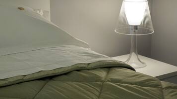 Een netjes opgemaakt bed met een groene deken en een wit kussen, een heldere glazen lamp op een witte nachtkastje, en een effen muur op de achtergrond.