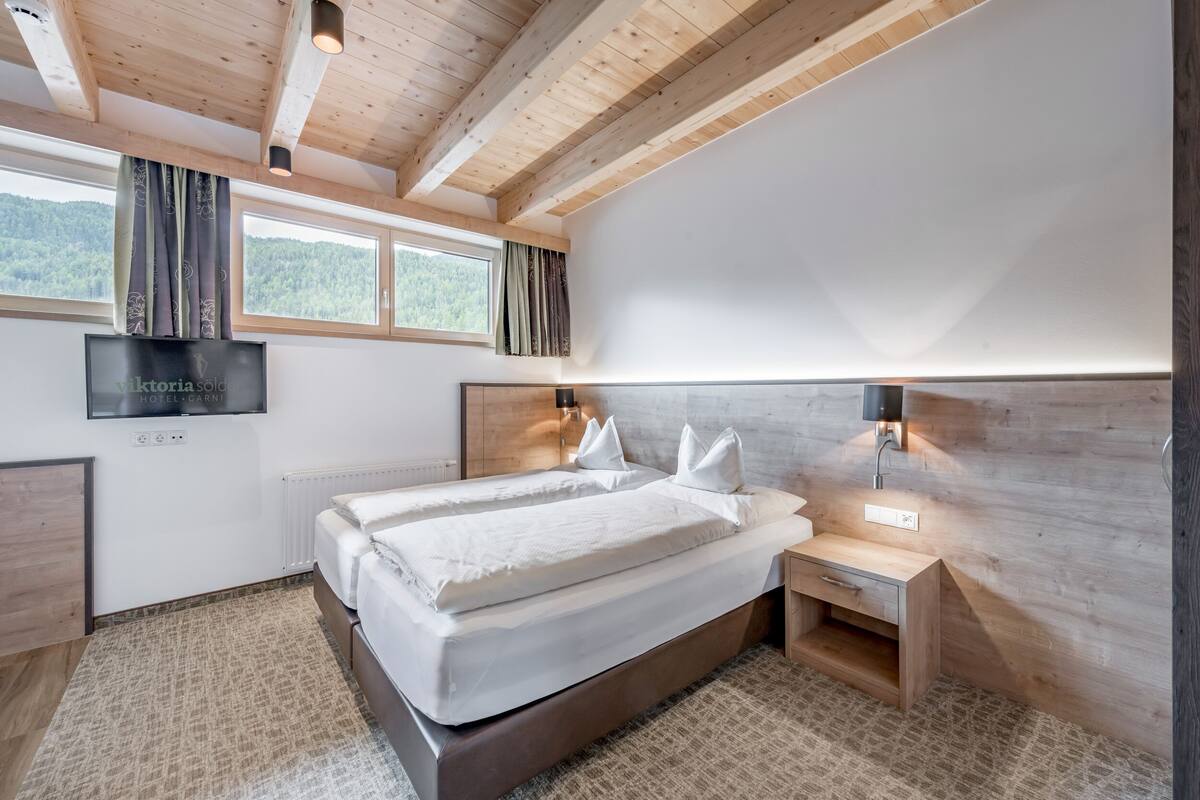A modern hotel room with a wooden ceiling, a large bed with white linens, a TV mounted on the wall, and a view of the outdoors through the window.
