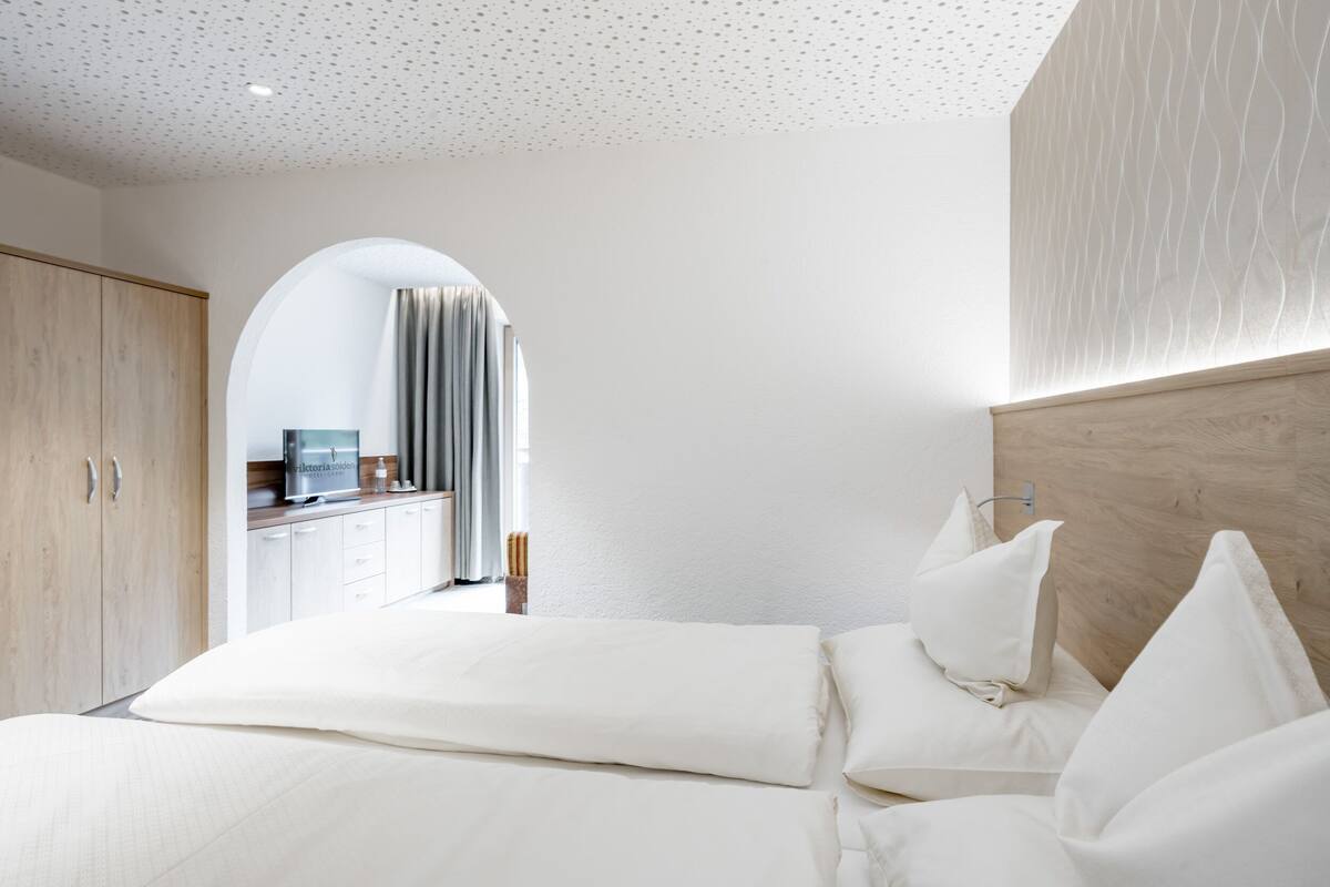 A modern hotel room with a large bed, wooden wardrobe, and a view through an arched doorway to a bathroom.