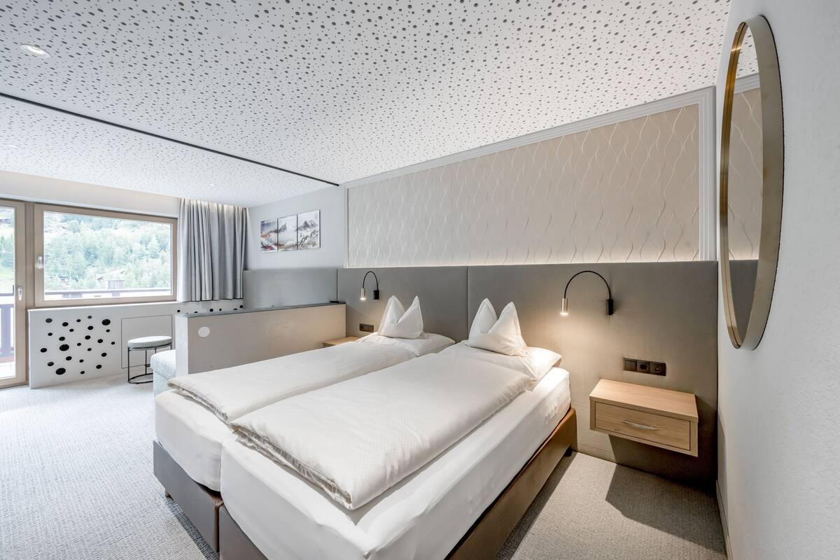 A modern bedroom with two beds, a large window, a wall-mounted mirror, and a small bedside table.