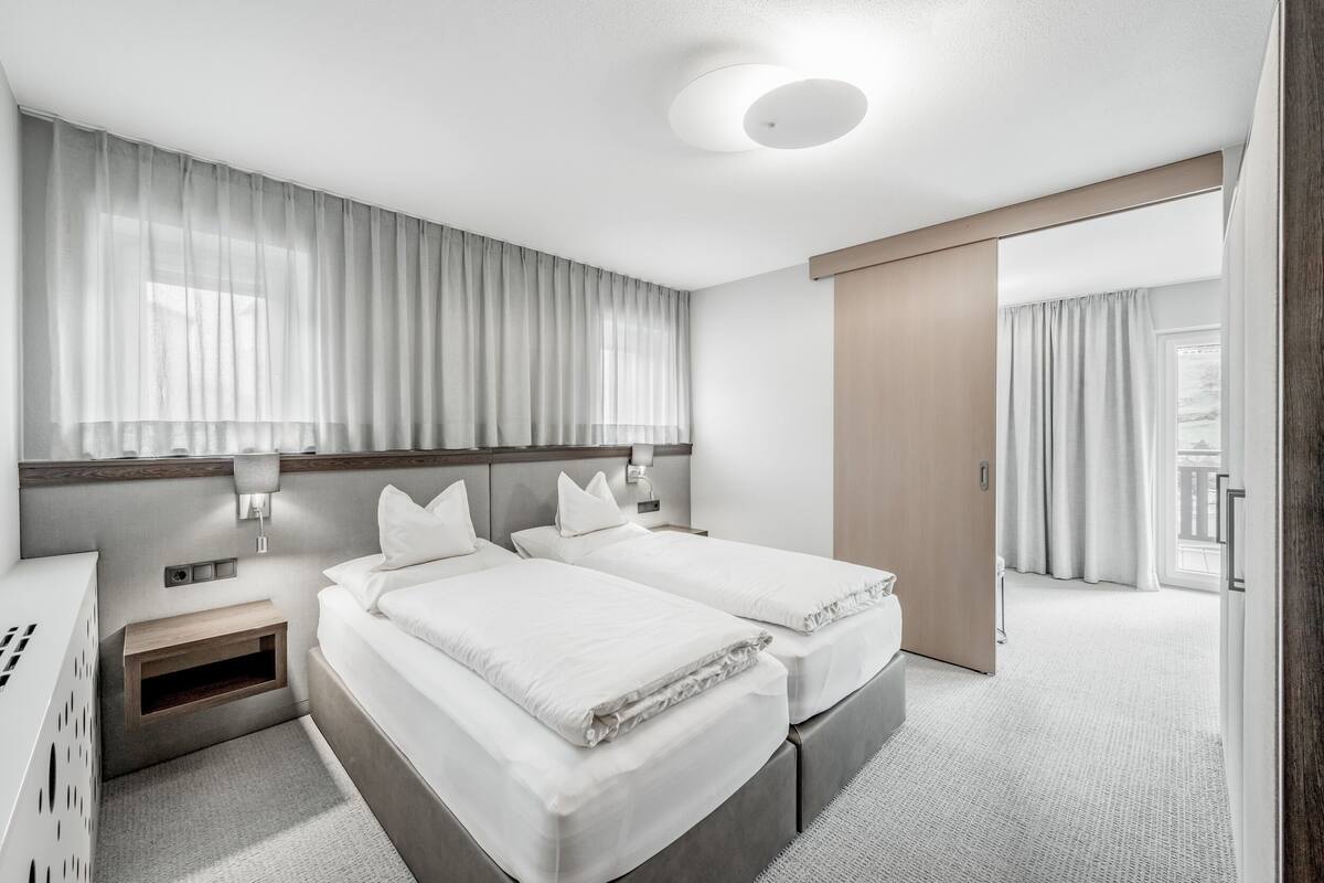 A modern hotel room with two beds, a bedside table, a nightstand, and a large window with sheer curtains.