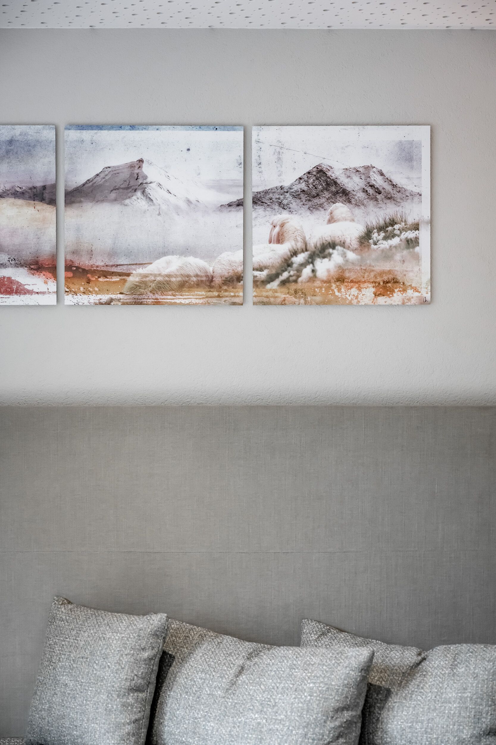 A room with a grey headboard, a grey sofa with patterned cushions, and a wall-mounted triptych painting depicting a snowy landscape.
