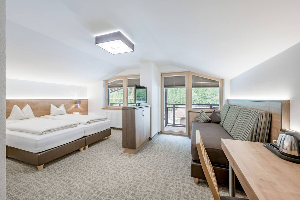 A modern hotel room with a large bed, a sofa, a desk, and a view of the outdoors.