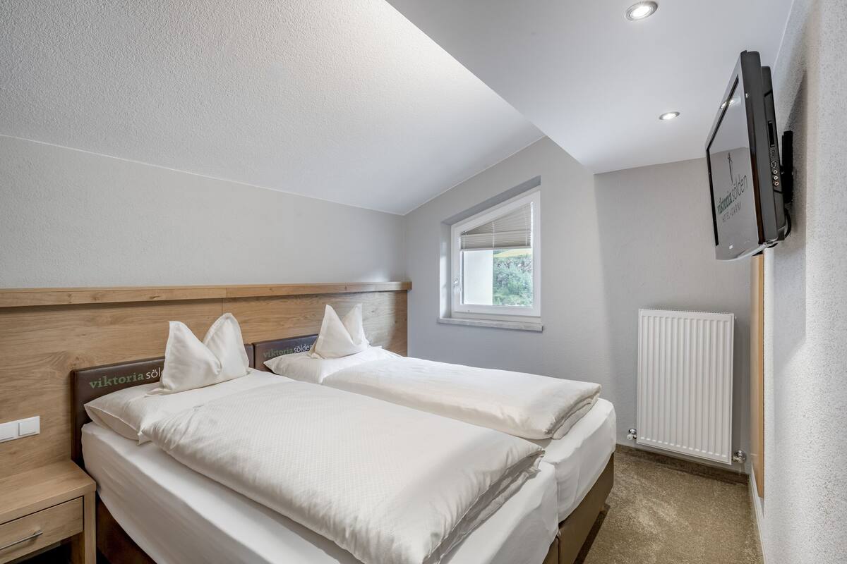 A bedroom with two beds, a wooden headboard, a window with blinds, a TV mounted on the wall, and a radiator.