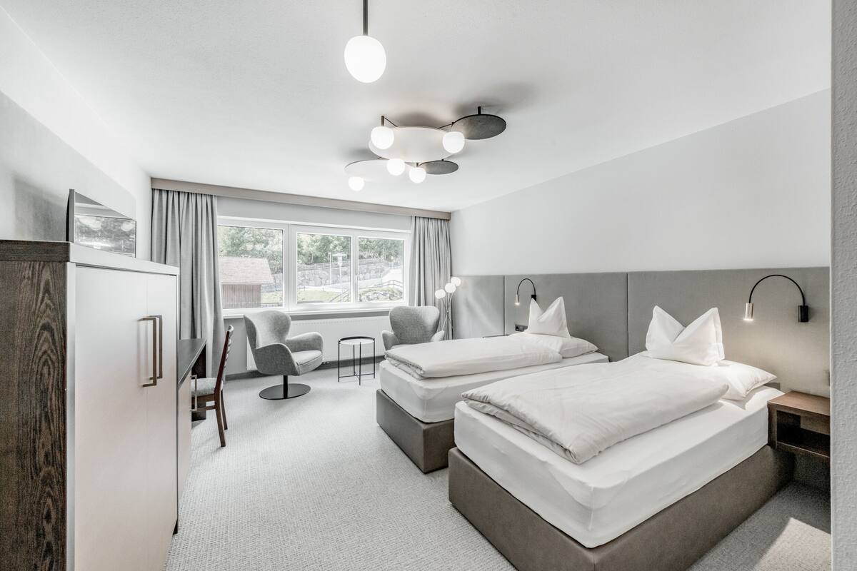 A modern hotel room with two beds, a desk, a chair, and a large window.