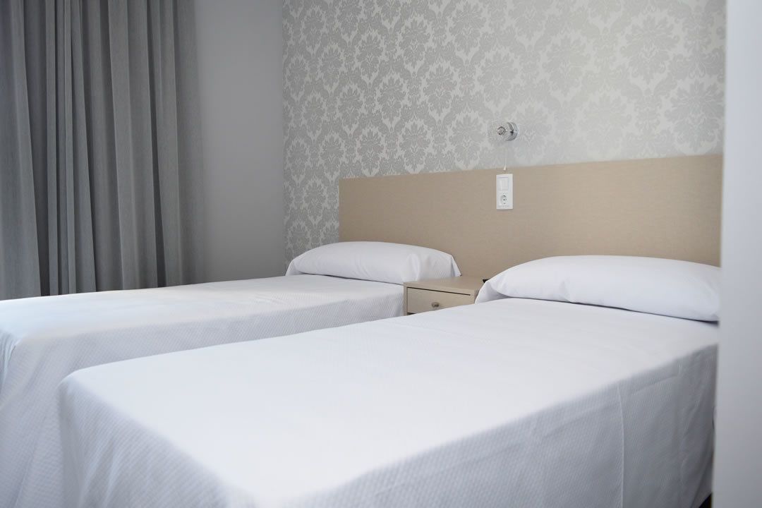 Habitación de hotel con dos camas individuales, una mesita de noche, un enchufe en la pared y una ventana con cortinas.
