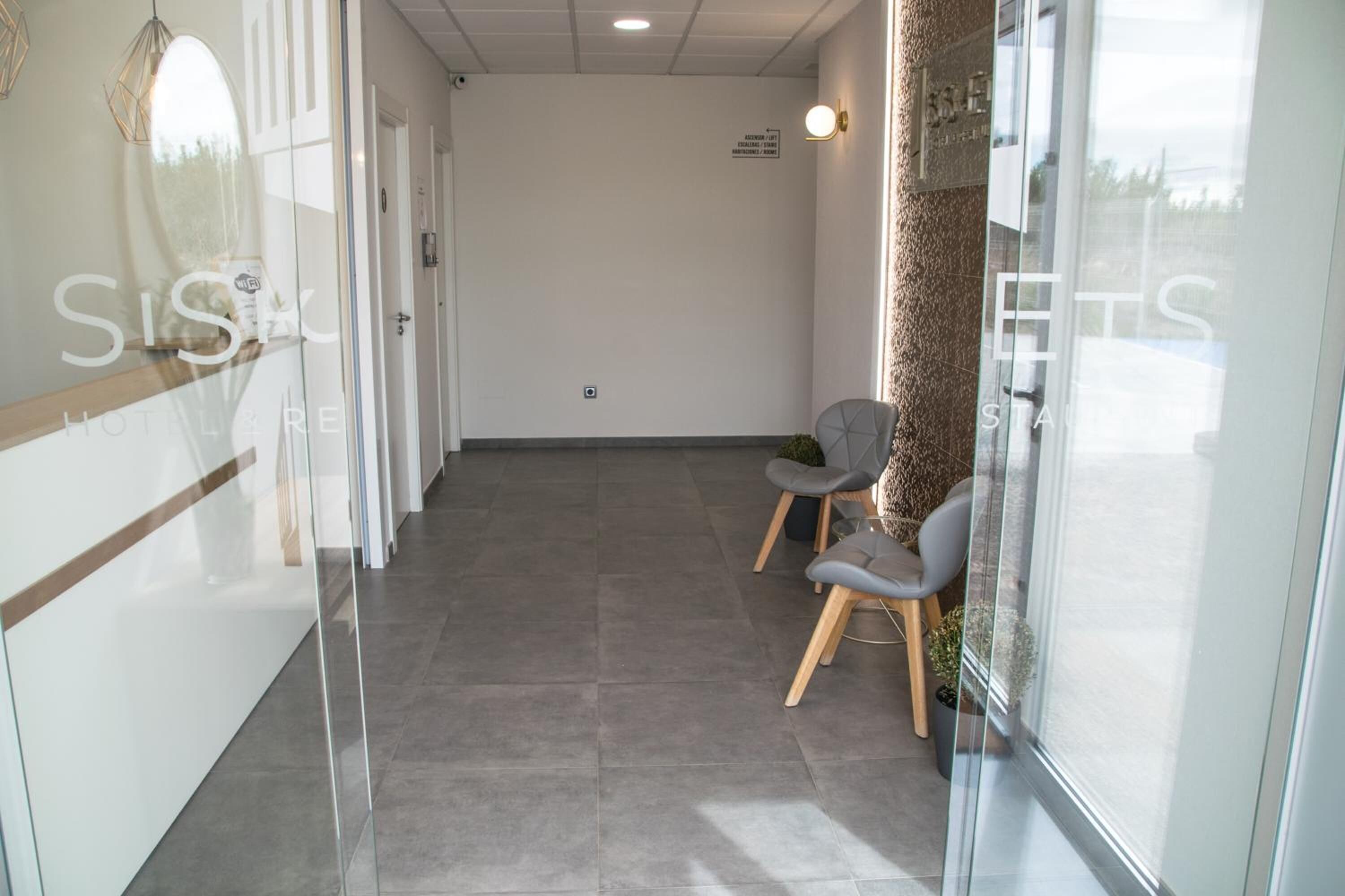 A hotel reception area with a glass wall, two grey chairs, a wooden bench, and a small potted plant.