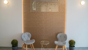 Een receptiegedeelte met een gestructureerde wand, twee grijze stoelen, een kleine ronde tafel en twee potplanten. Een bord boven de stoelen geeft 'Sikets Hotel & Restaurant' weer.
