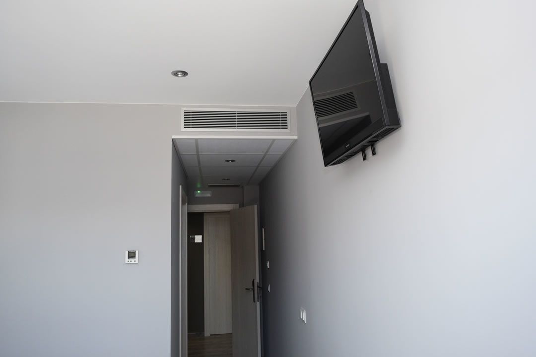 Un couloir avec un téléviseur à écran plat fixé au mur, une bouche d’aération au plafond et une porte donnant sur une autre pièce.