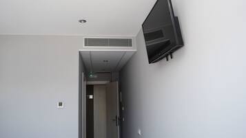 Un couloir avec un téléviseur à écran plat fixé au mur, une bouche d’aération au plafond et une porte donnant sur une autre pièce.
