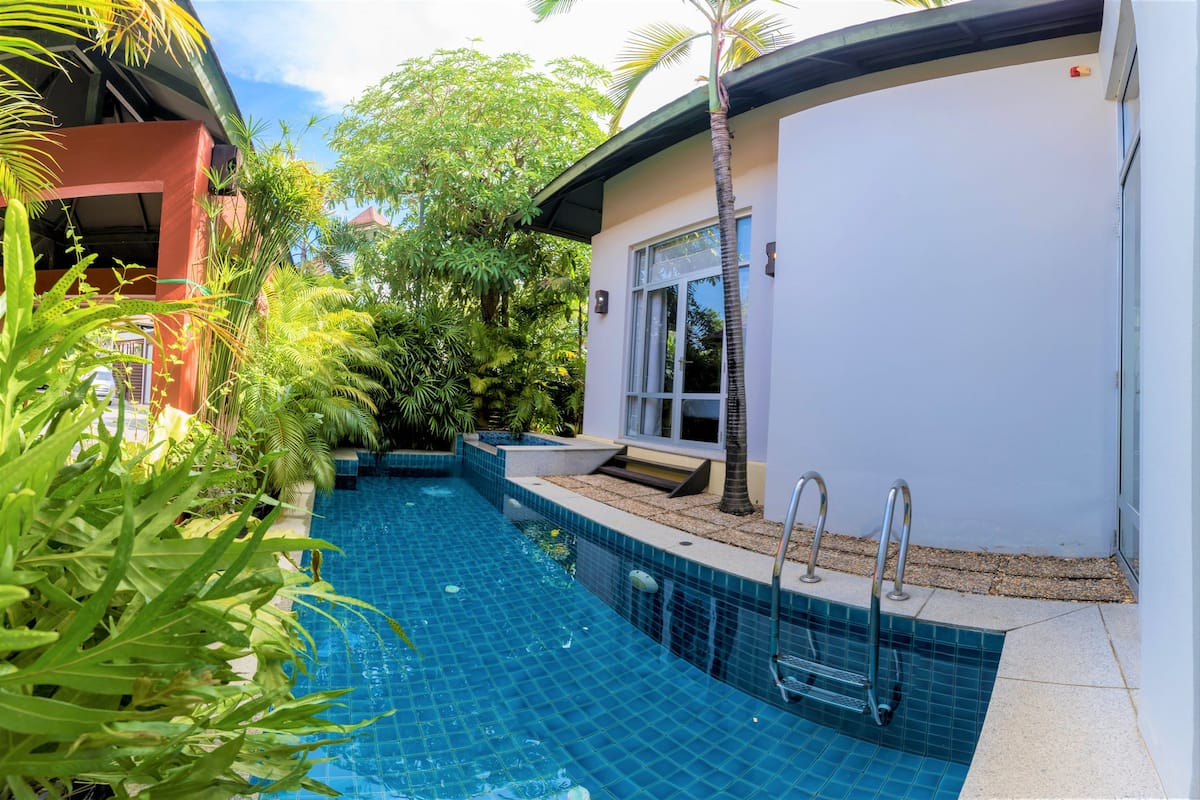 A swimming pool with a tiled edge, surrounded by lush greenery and a white building with a glass door.