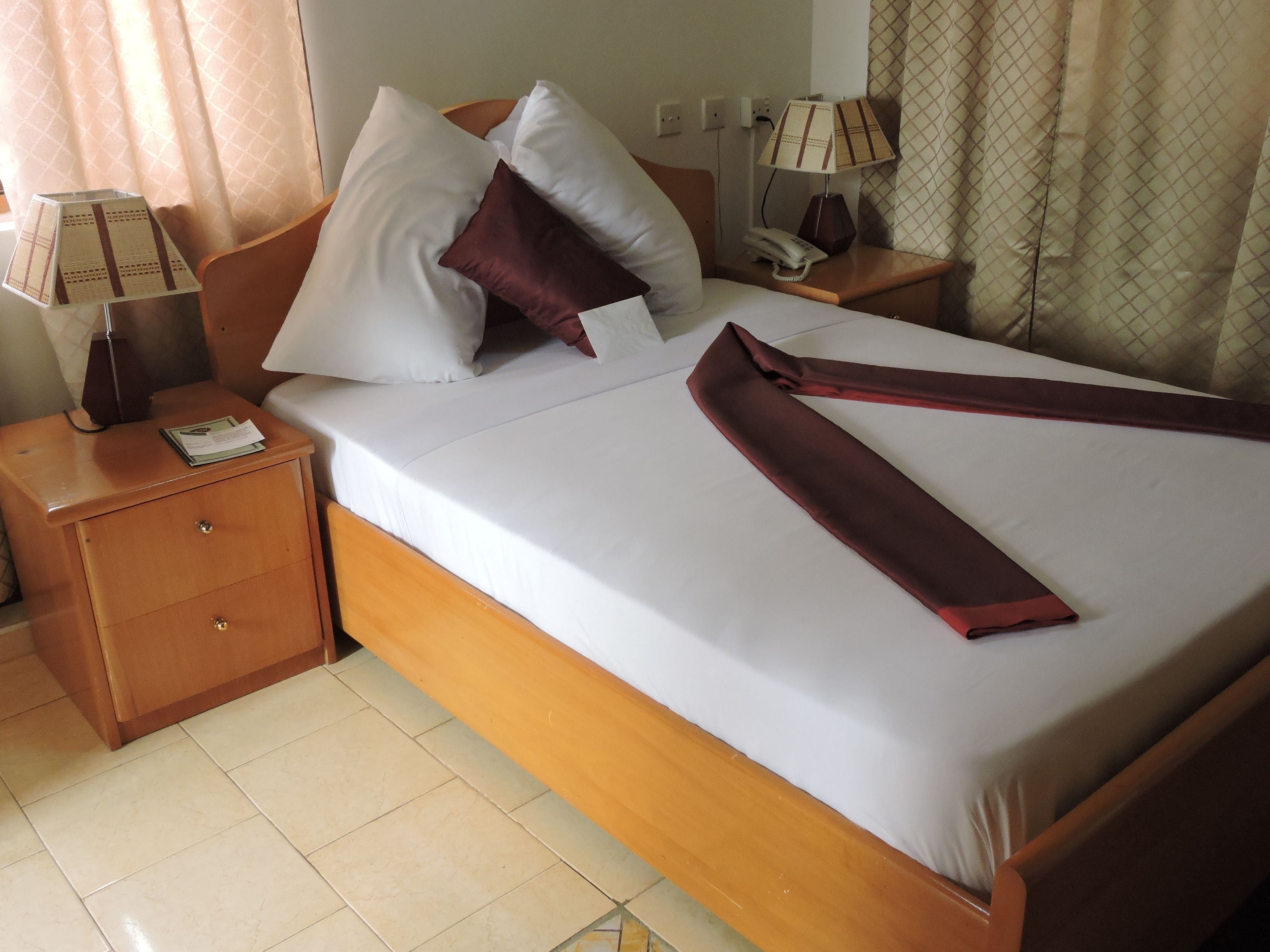 A hotel room with a neatly made bed, a wooden nightstand with a lamp, a telephone, and a drawer.
