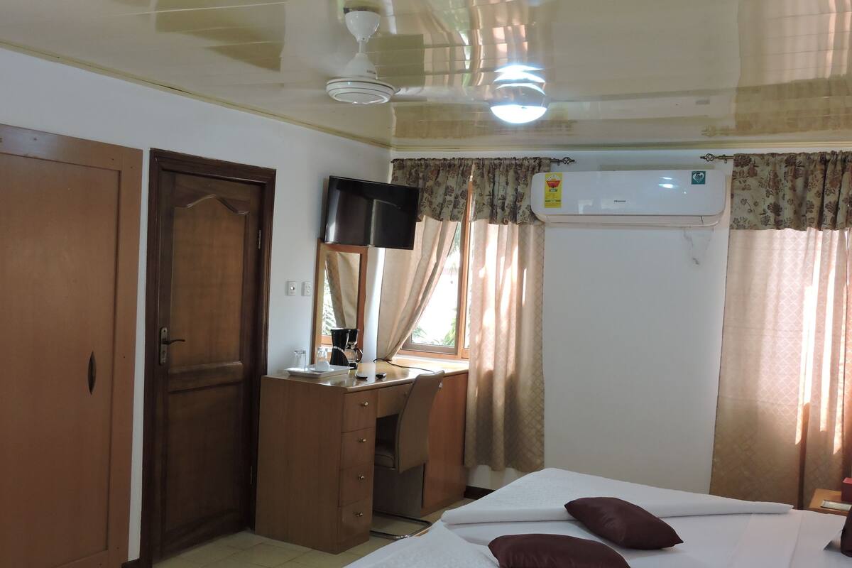 A hotel room with a bed, a desk with a mirror, a ceiling fan, and an air conditioning unit.