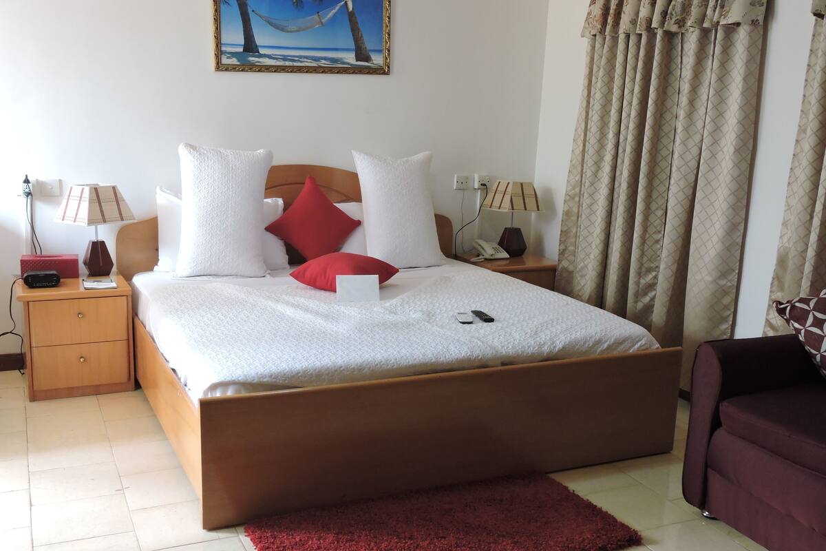 A neatly made bed with white bedding and red pillows, a wooden nightstand with a lamp, a telephone, and a small table with a lamp.