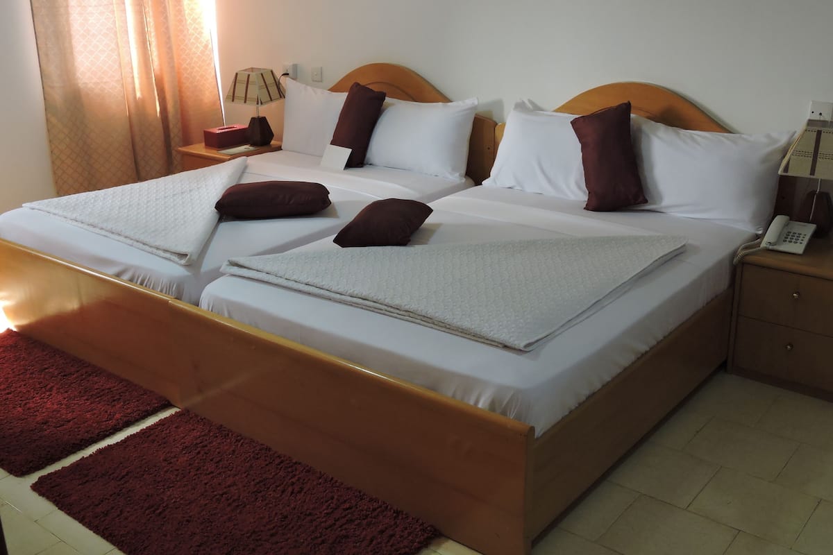 A neatly made bed with white linens and brown pillows, a wooden headboard, a red rug, and a bedside table with a lamp and a phone.