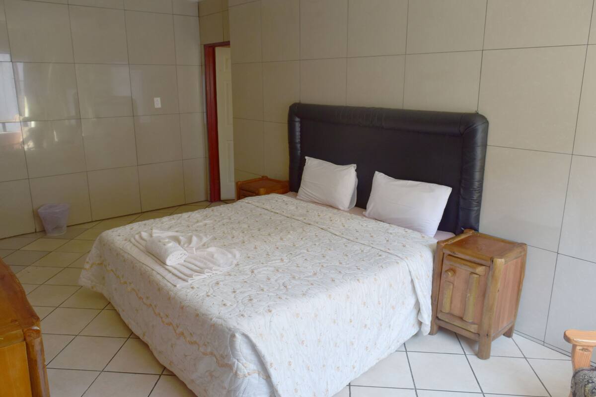 A neatly made bed with a white bedspread and two pillows, a wooden nightstand with a drawer, and a tiled floor.