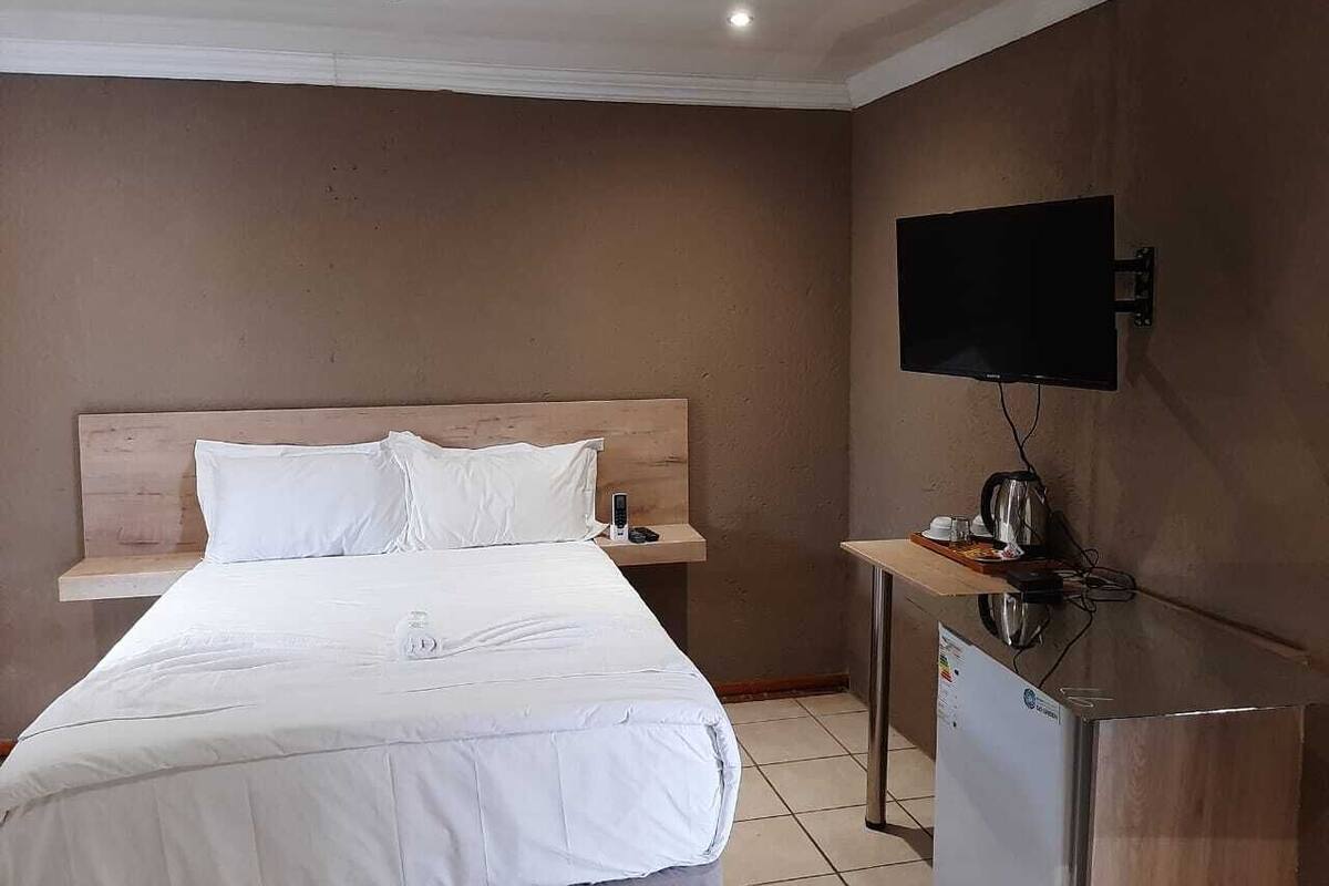 A hotel room with a bed, a bedside table with a kettle, a TV mounted on the wall, and a small refrigerator.
