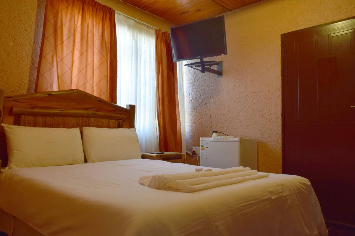 A hotel room with a wooden headboard, a flat-screen TV mounted on the wall, a bedside table with a remote control, and a small refrigerator.