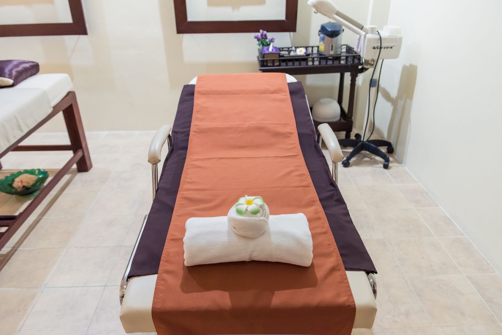 Une table de massage recouverte d’une serviette marron et agrémentée d’un petit objet décoratif, un banc en bois avec un coussin, et une petite table sur laquelle se trouvent un vase avec une fleur et une tasse.