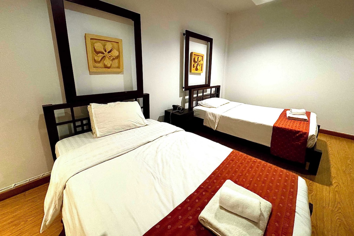 A hotel room with two single beds, each with a white bedspread and a red blanket. There are framed pictures on the wall above each bed.