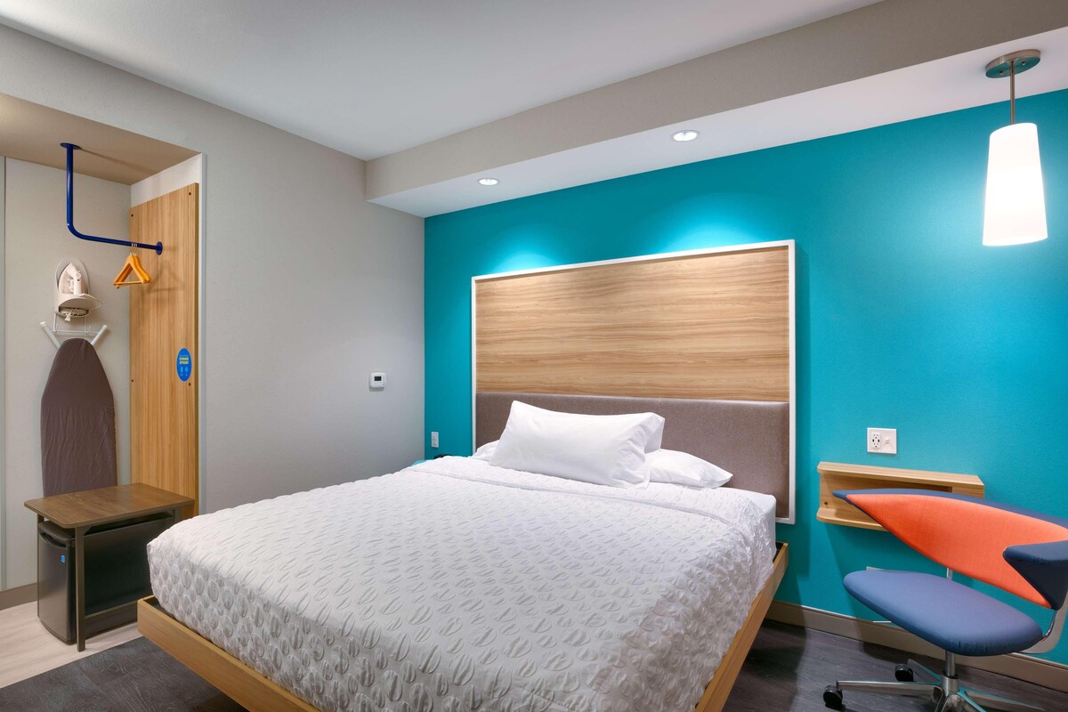 A modern hotel room with a bed, a chair, a desk, and a wardrobe.