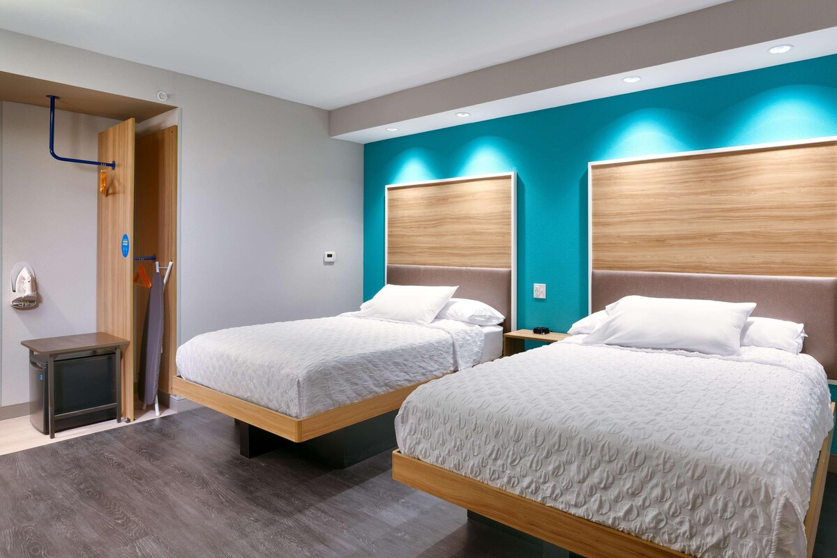 A hotel room with two beds, wooden headboards, a small bedside table, a wall-mounted heater, and a door with a towel rack.